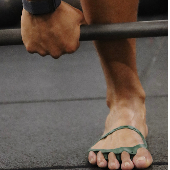 Person wearing green sandals lifting a barbell on a gym floor.